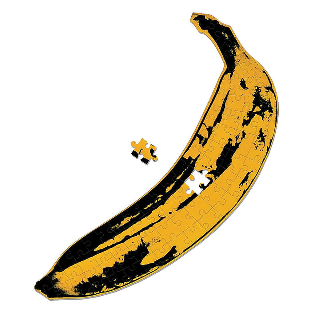 A close-up view of the Andy Warhol Banana Mini Shaped Puzzle by Galison on a white background, showing the complete puzzle and a single loose piece near the packaging. Great for fans of Andy Warhol art and collectible jigsaw puzzles.